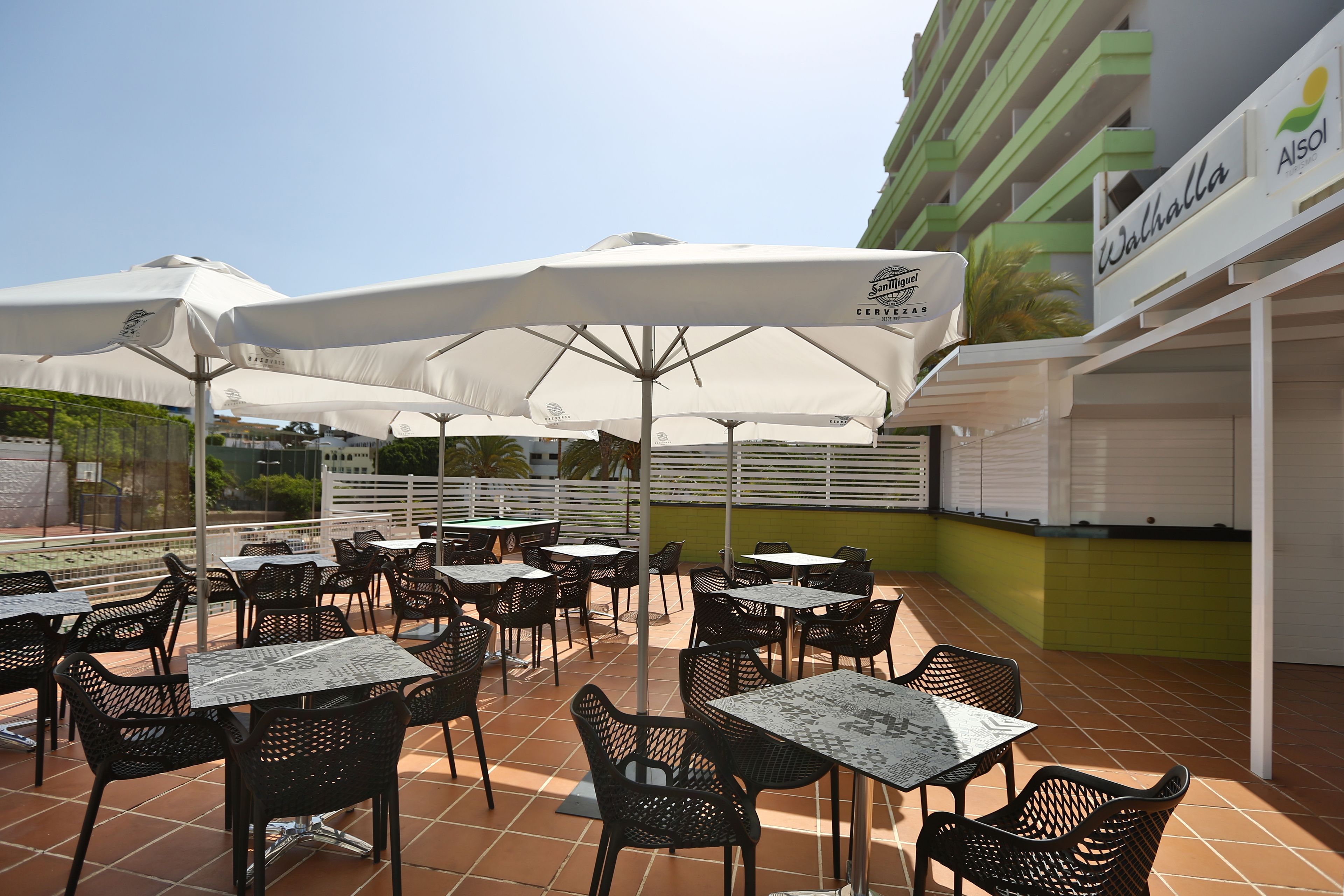 An outdoor patio area with multiple tables and chairs, some under white umbrellas, and a building with a green and white facade in the background.