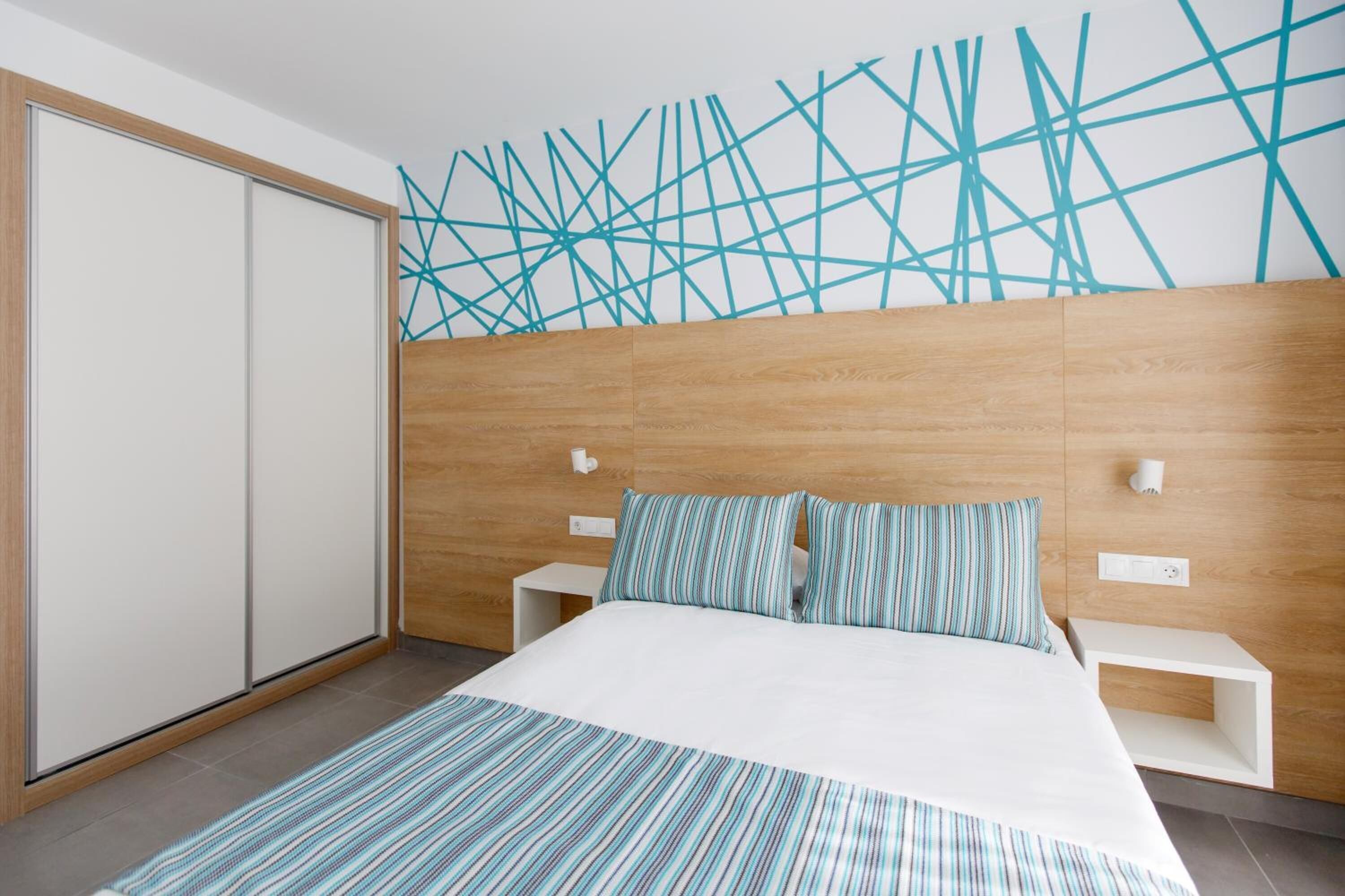 A modern bedroom with a wooden headboard, a large bed with striped bedding, two bedside tables, and a geometric-patterned wall.
