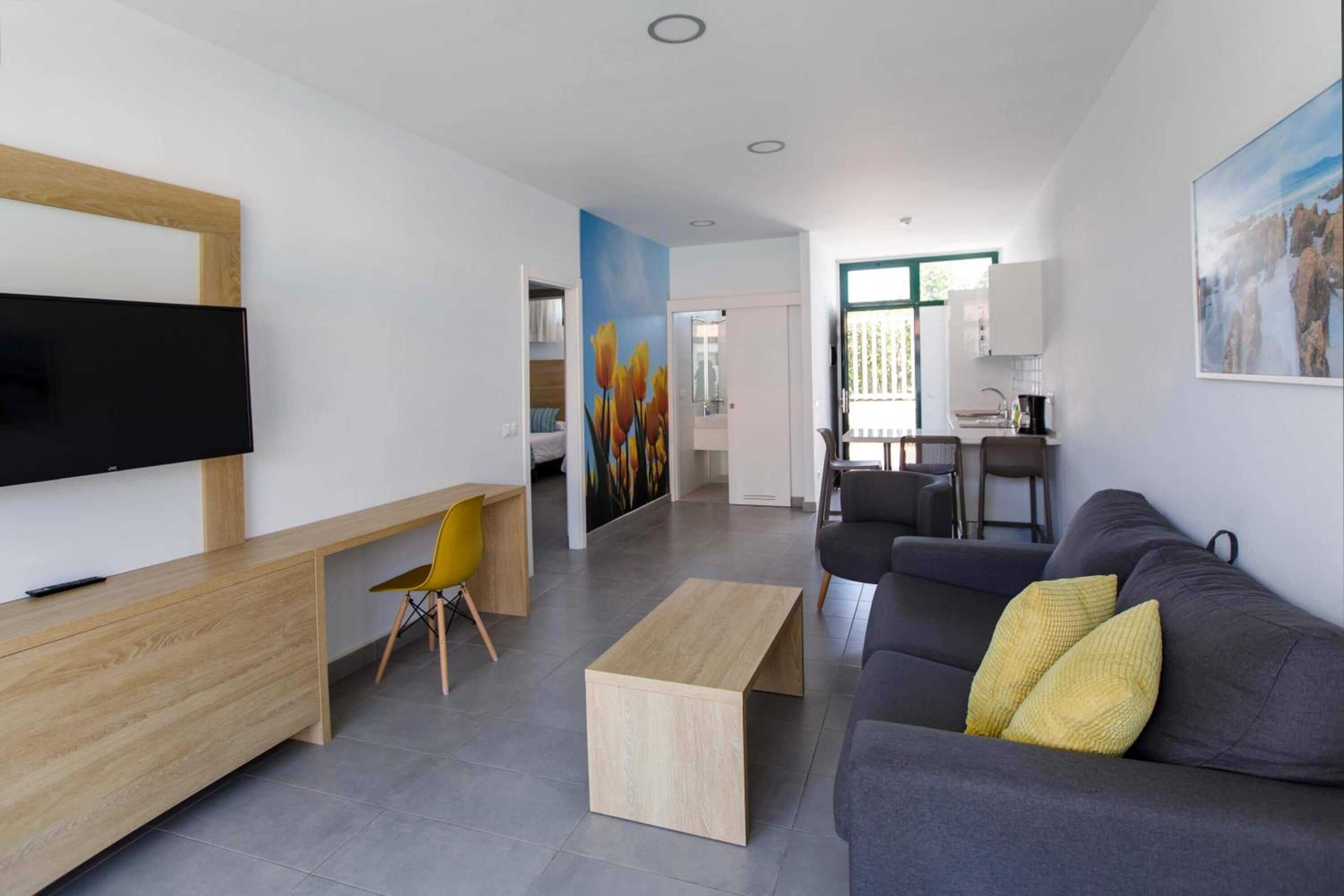 A modern living room with a flat-screen TV, a wooden desk, a grey sofa with yellow cushions, and a painting on the wall.