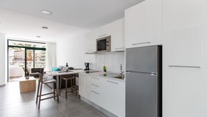 A modern kitchen with white cabinets, a stainless steel refrigerator, and a microwave. There is a dining area with a table and chairs, and a window providing a view of palm trees.