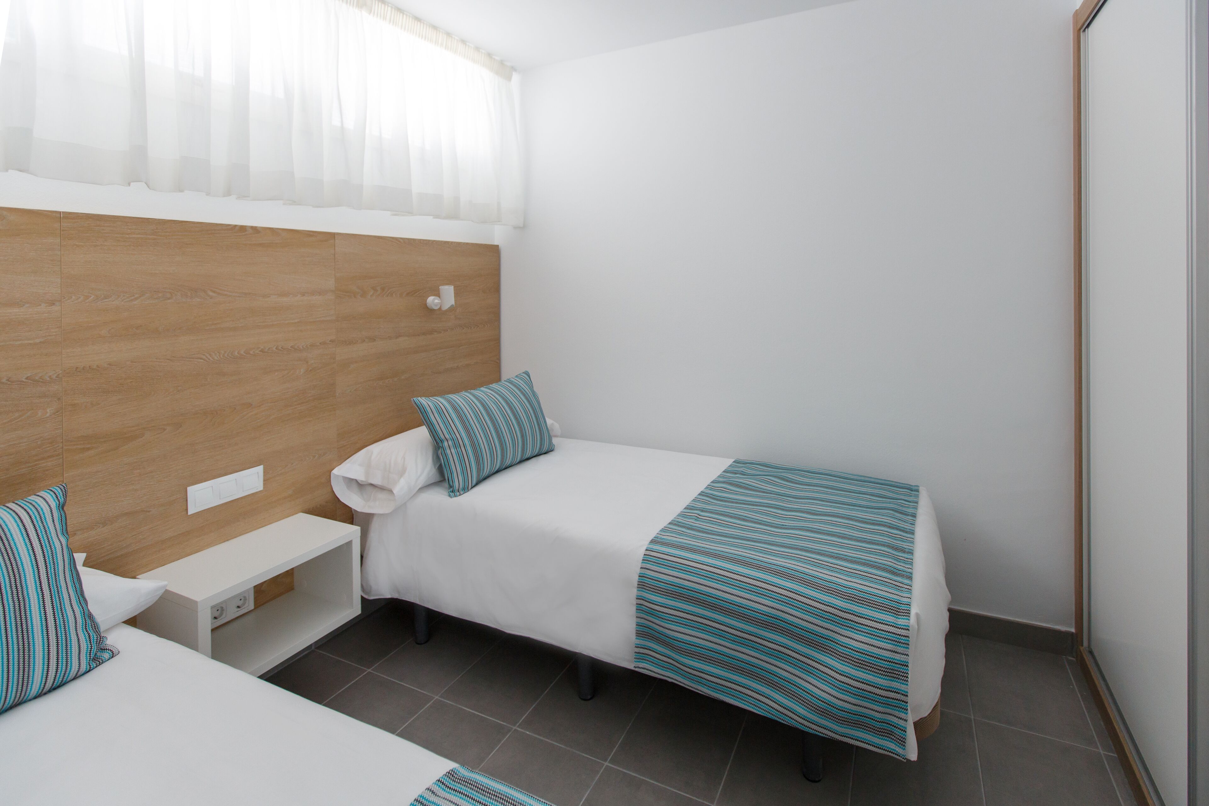 A bedroom with two single beds, a wooden headboard, a small bedside table, and a window with sheer curtains.