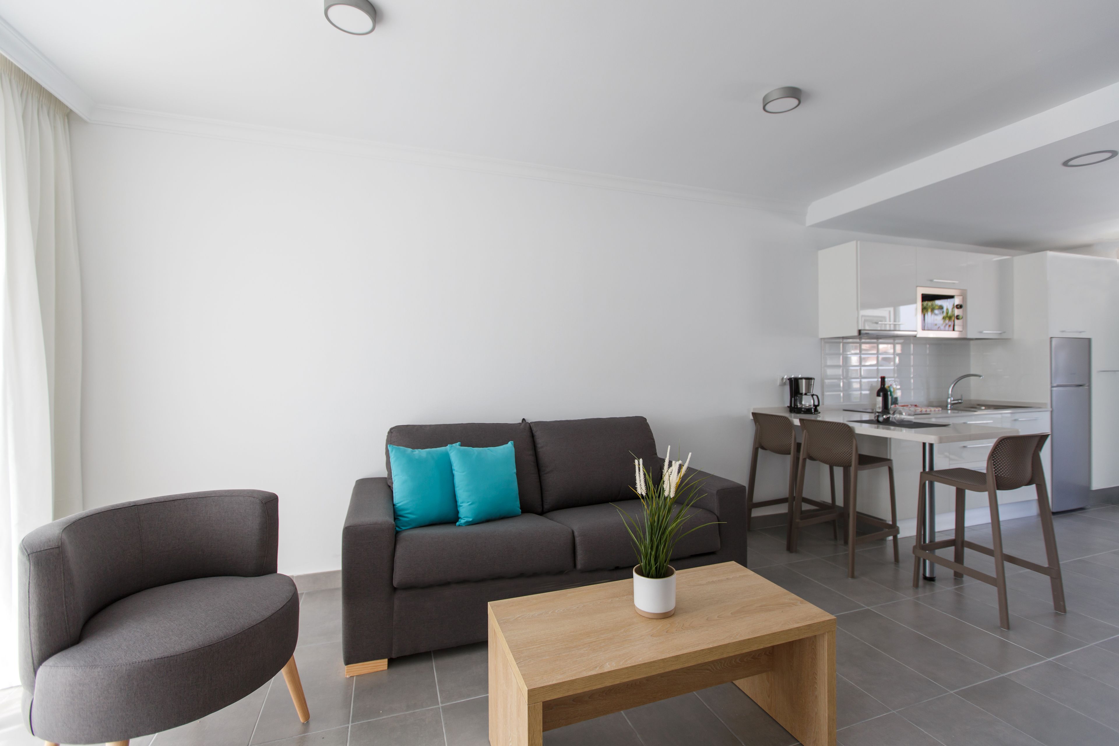 A modern living room with a grey sofa, a wooden coffee table, and a kitchen area with a white countertop and bar stools.