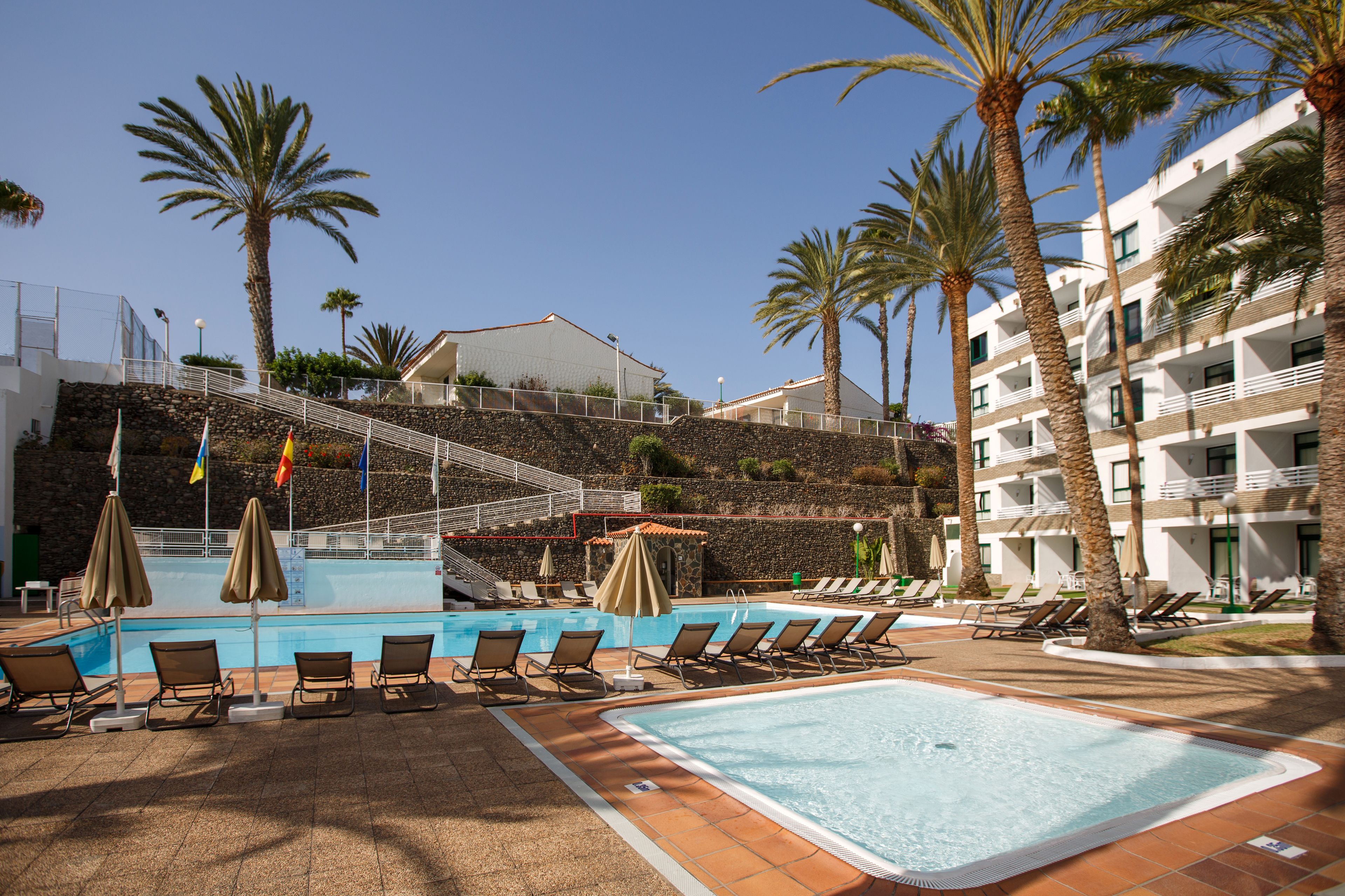 A pool area with lounge chairs and umbrellas, a stone wall, and a building in the background.