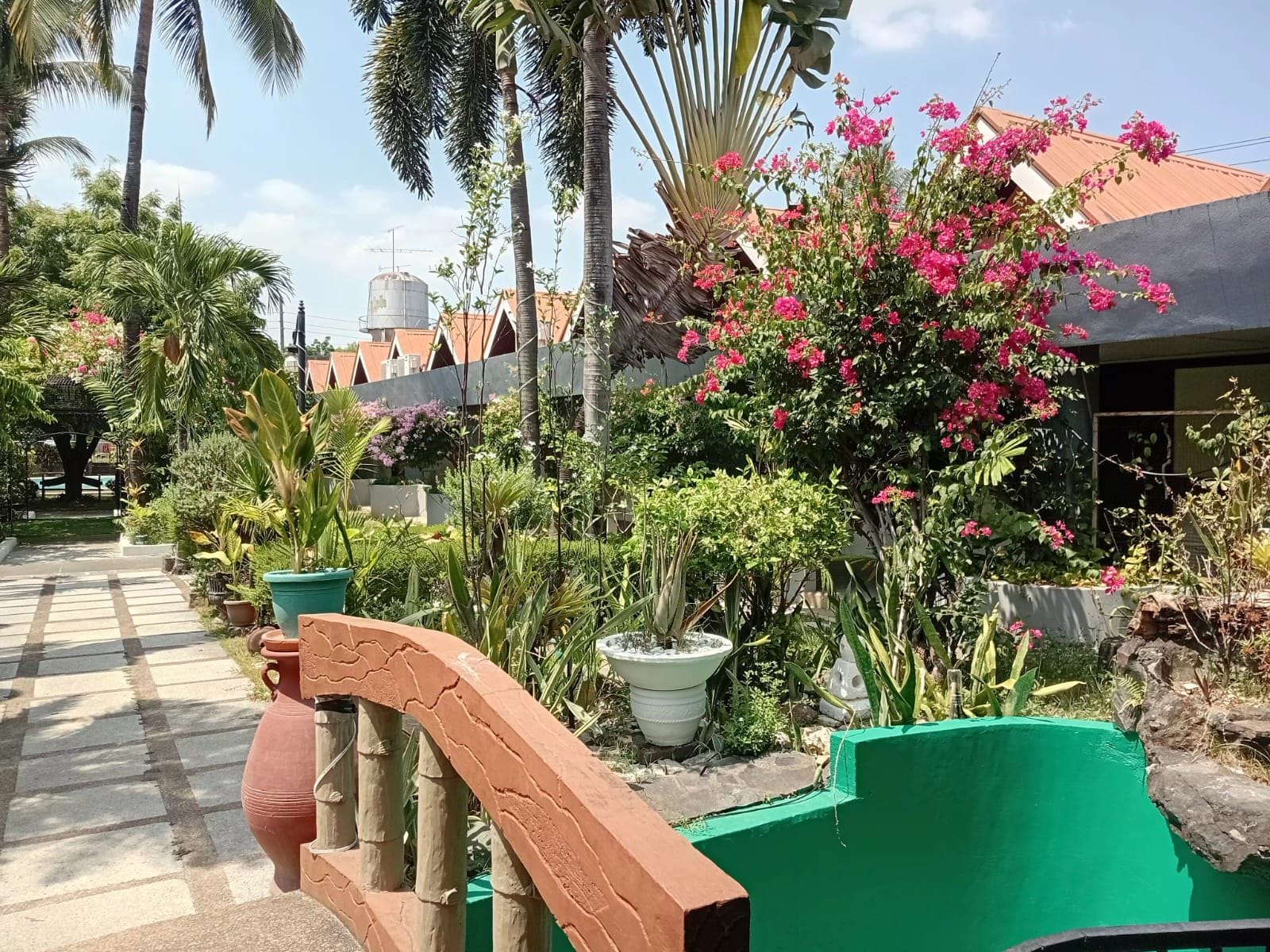 Un camino lleva a un edificio con un depósito de agua, rodeado de vegetación exuberante y plantas florecientes.
