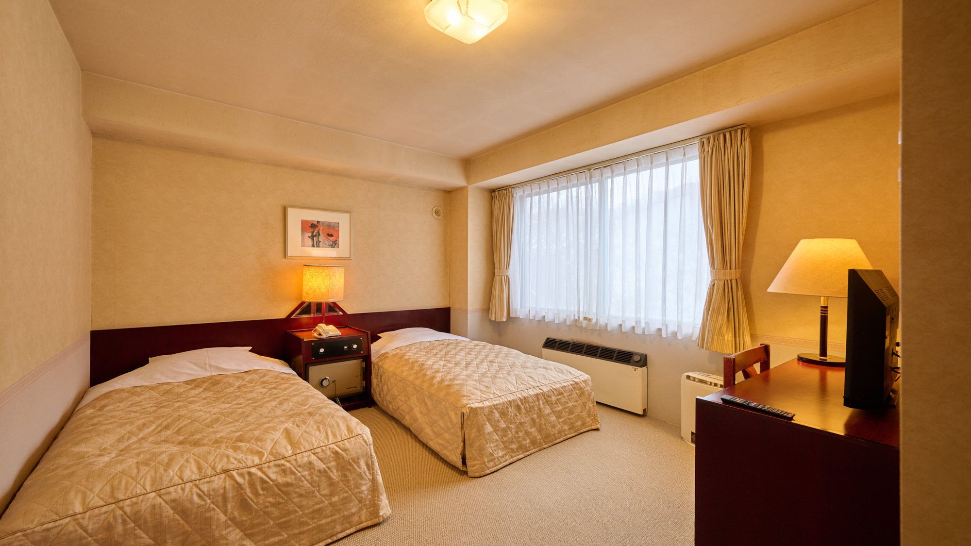 Una habitación de hotel con dos camas, un escritorio con lámpara, un calentador y una ventana con cortinas.