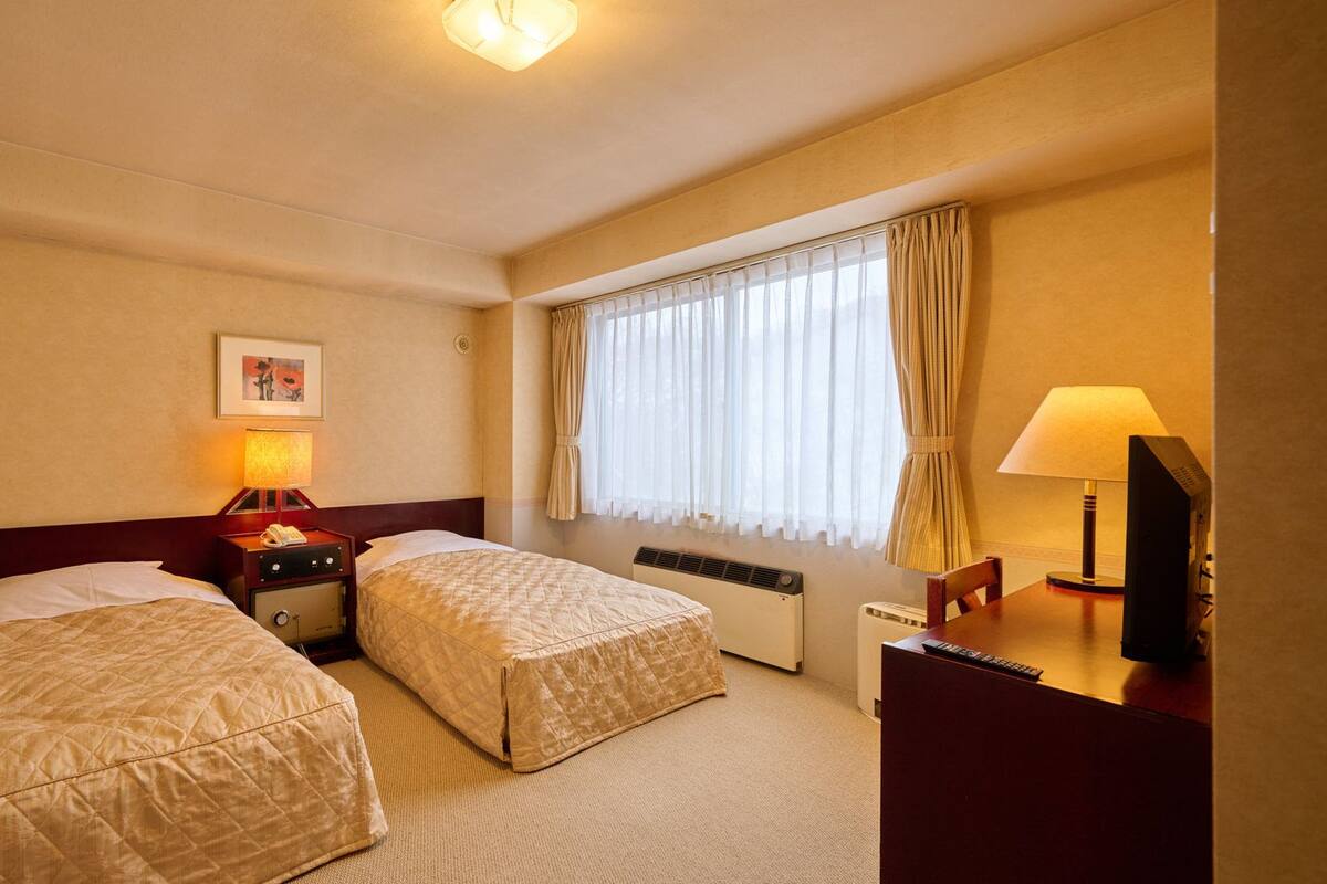 A hotel room with two beds, a desk with a lamp, a heater, and a window with curtains.