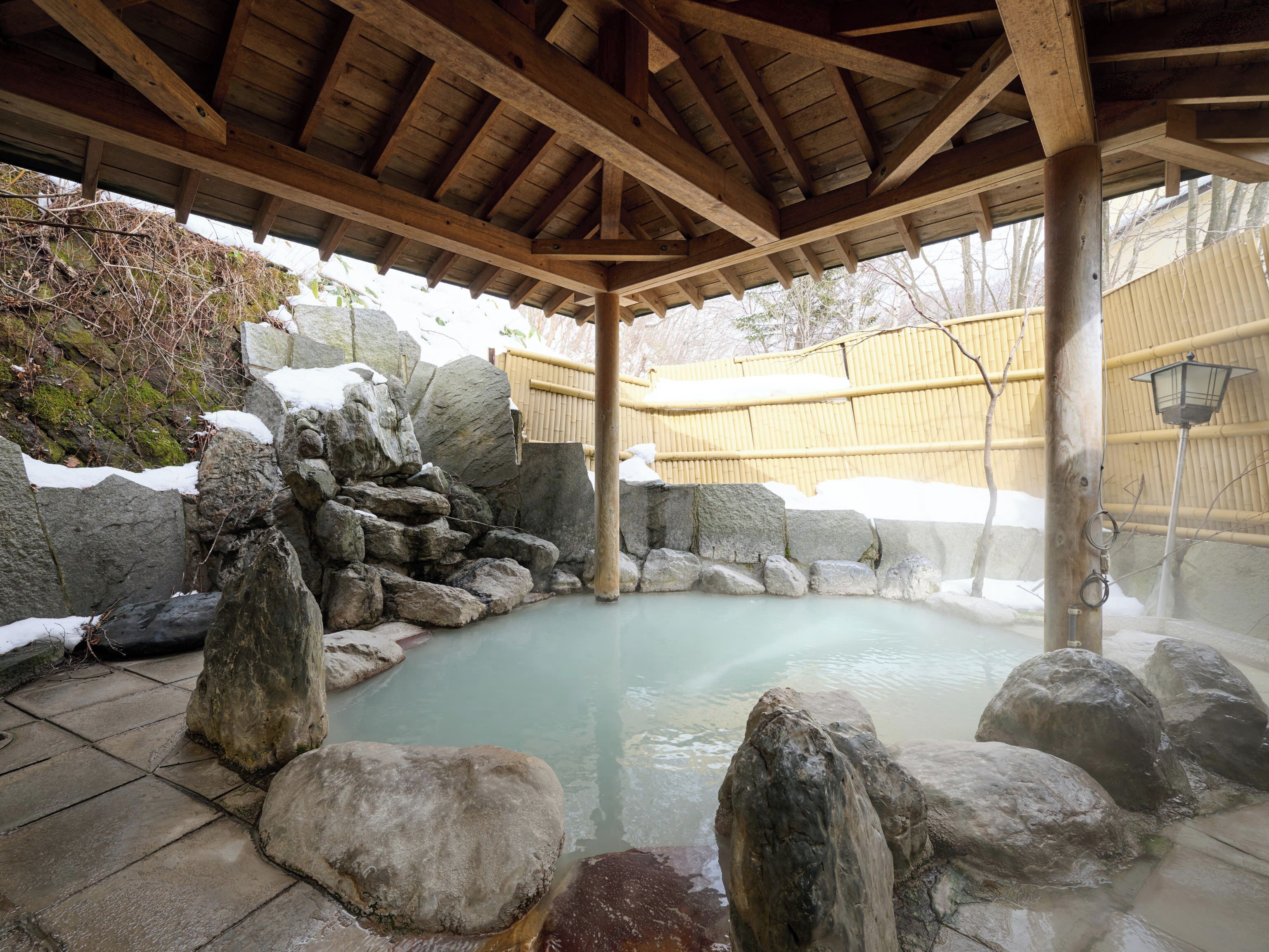 A stone hot spring surrounded by rocks, wooden beams, and a bamboo fence.