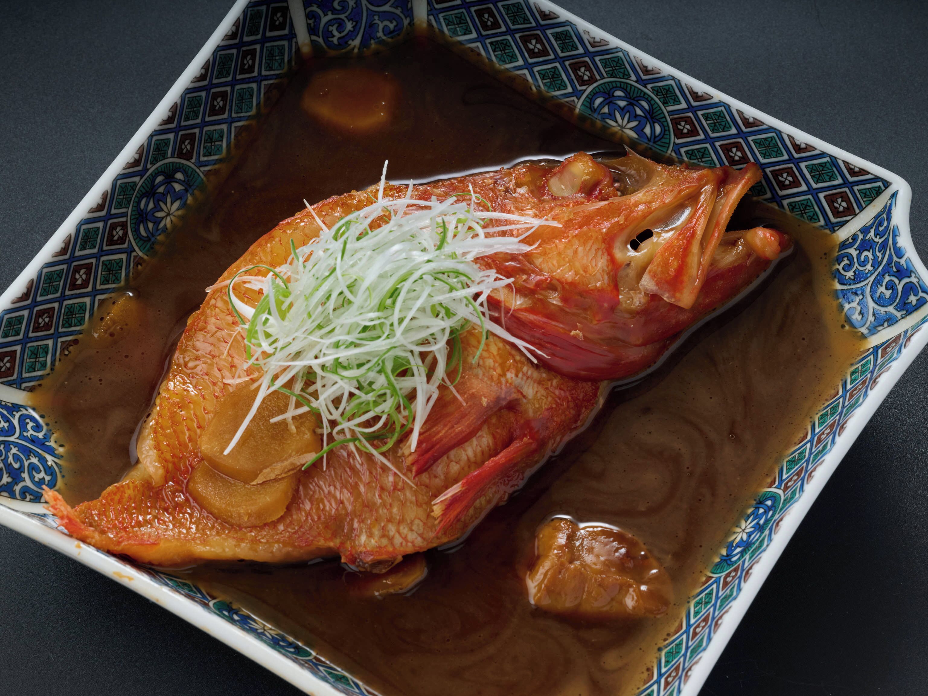 A square plate with a patterned border containing a whole fish topped with shredded green vegetables and a brown sauce.