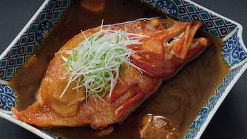 A square plate with a patterned border containing a whole fish topped with shredded green vegetables and a brown sauce.