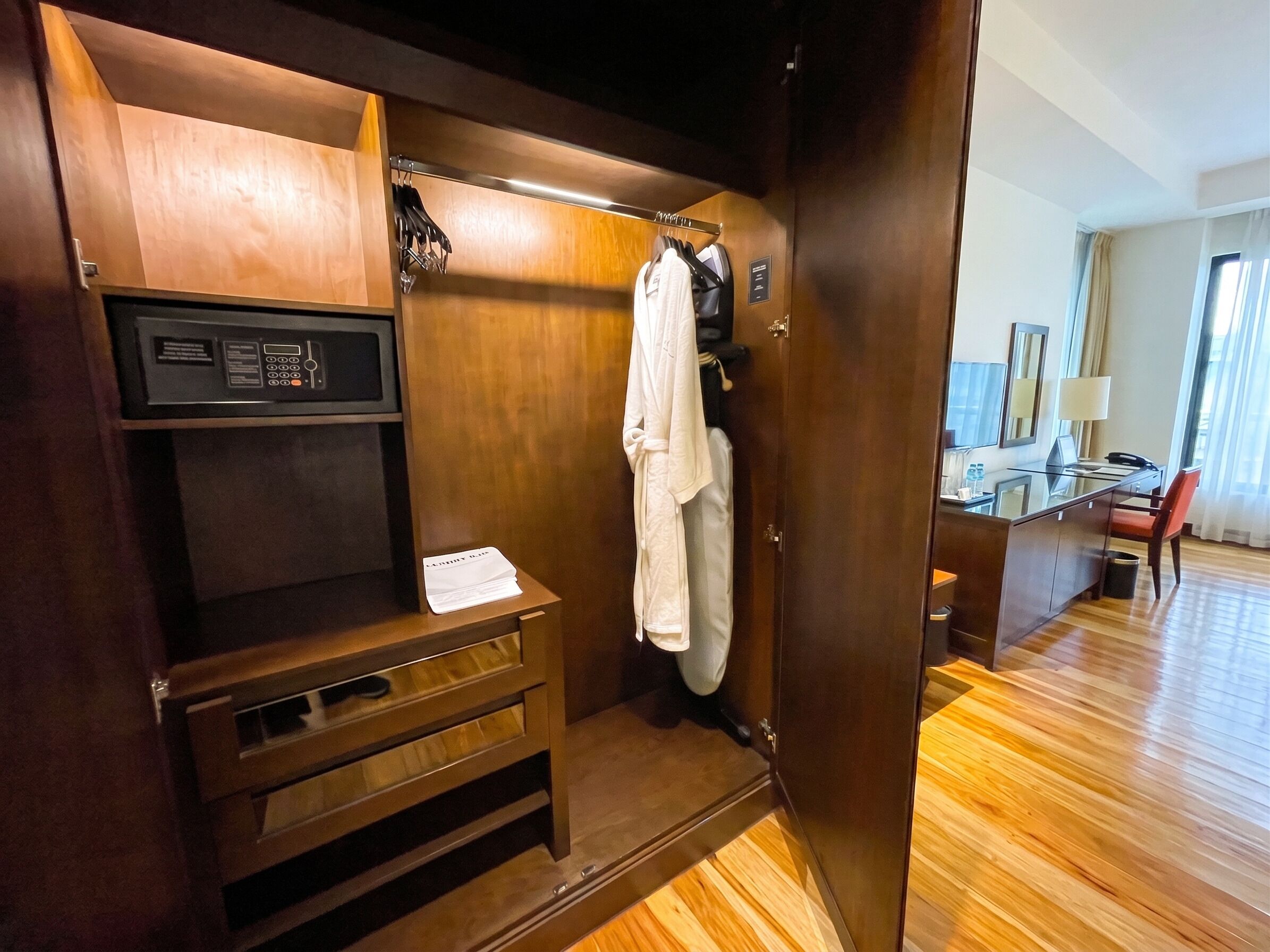 Habitación de hotel con un armario de madera, un escritorio y una silla, un espejo y una ventana con cortinas.