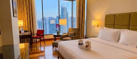 A hotel room with a large bed, a desk with a chair, a small table with a lamp, and a view of the cityscape through large windows.