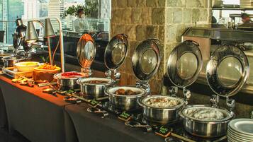 A buffet setup with various food items and utensils, including pots of rice and other dishes, in a hotel setting with a city view.