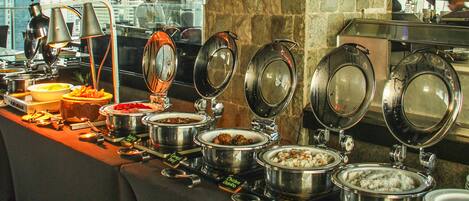 A buffet setup with various food items and utensils, including pots of rice and other dishes, in a hotel setting with a city view.