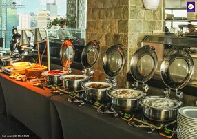 A buffet setup with various food items and utensils, including pots of rice and other dishes, in a hotel setting with a city view.
