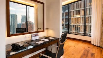 A hotel room with a wooden desk, a black office chair, a large mirror, a telephone, a calendar, and a view of tall buildings through the window.