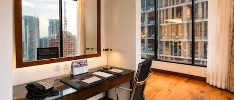 Habitación de hotel con un escritorio de madera, una silla de oficina negra, un espejo grande, un teléfono, un calendario y vistas de edificios altos a través del ventanal.