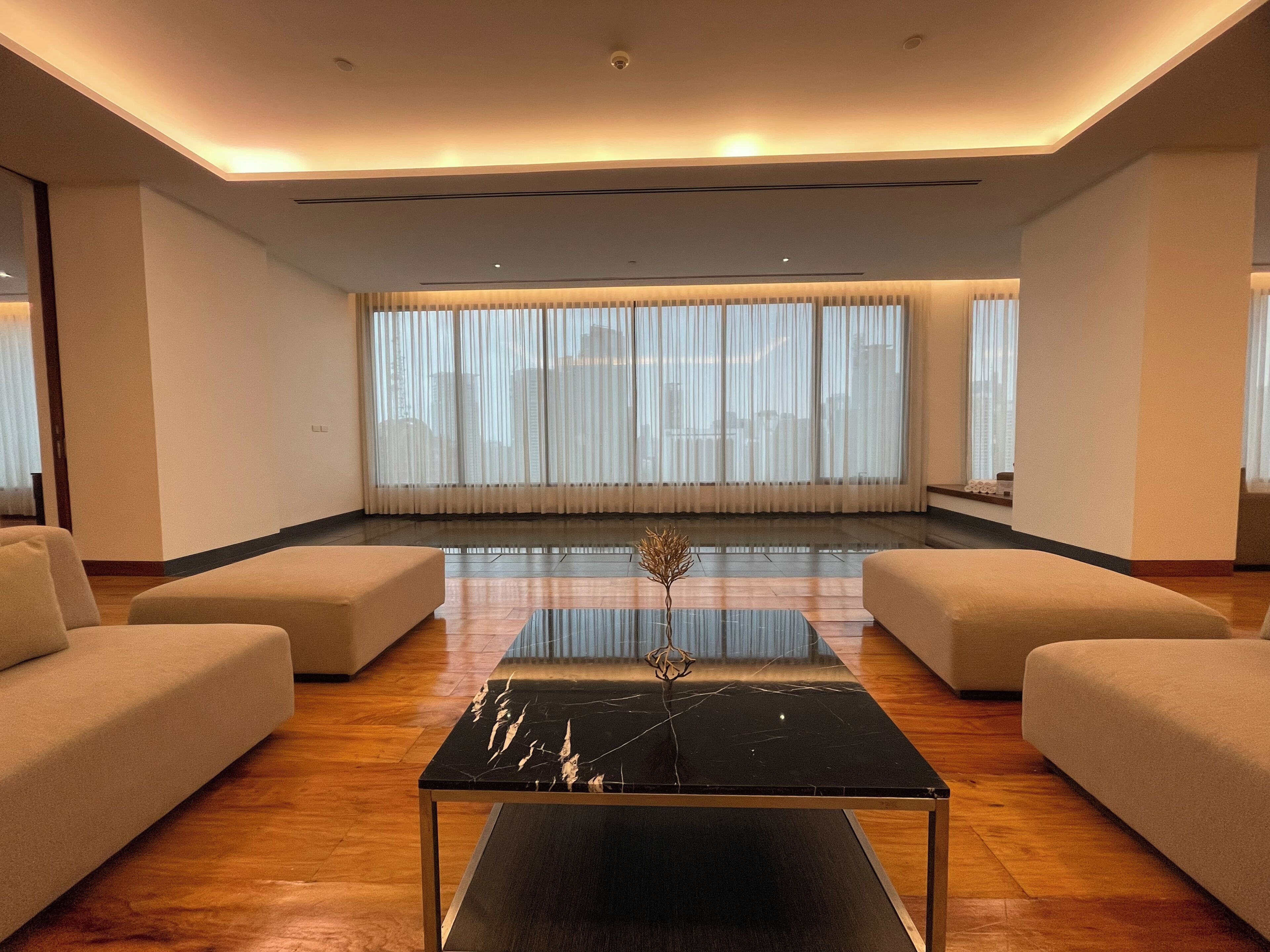 A modern living room with a black marble coffee table, beige sofas, and large windows with sheer curtains.