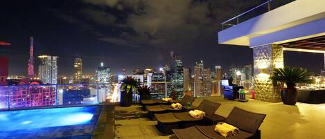 A rooftop pool area with lounge chairs and a city skyline view at night.
