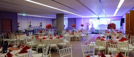 Un salón de banquetes con mesas redondas, sillas blancas y servilletas rojas.