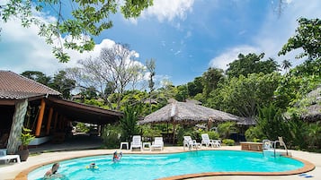 A swimming pool surrounded by lounge chairs and thatched umbrellas, with a tropical setting.