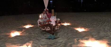 A boat with a canopy and a bow, decorated with a bow and a white ribbon, on a sandy beach with fires around it.