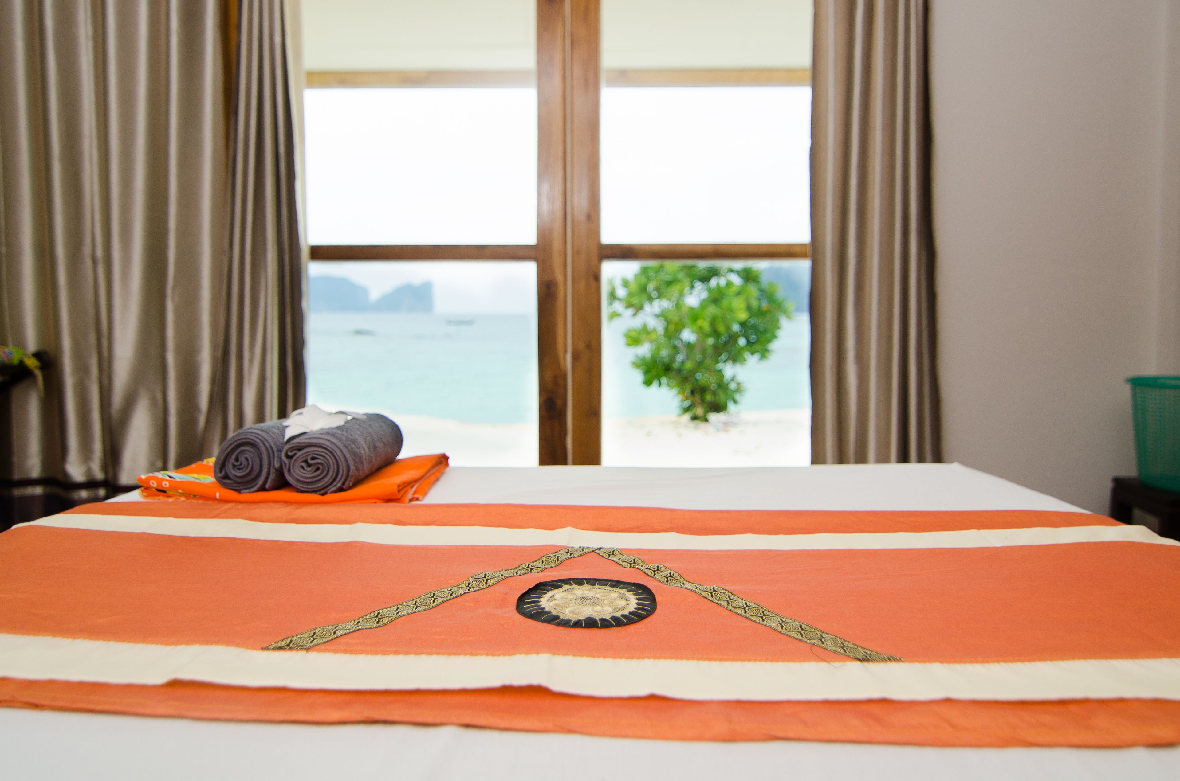 Un lit avec une couvre-lit orange et un coussin décoratif, une fenêtre donnant sur la mer et un arbre, et un rideau sur le côté gauche.