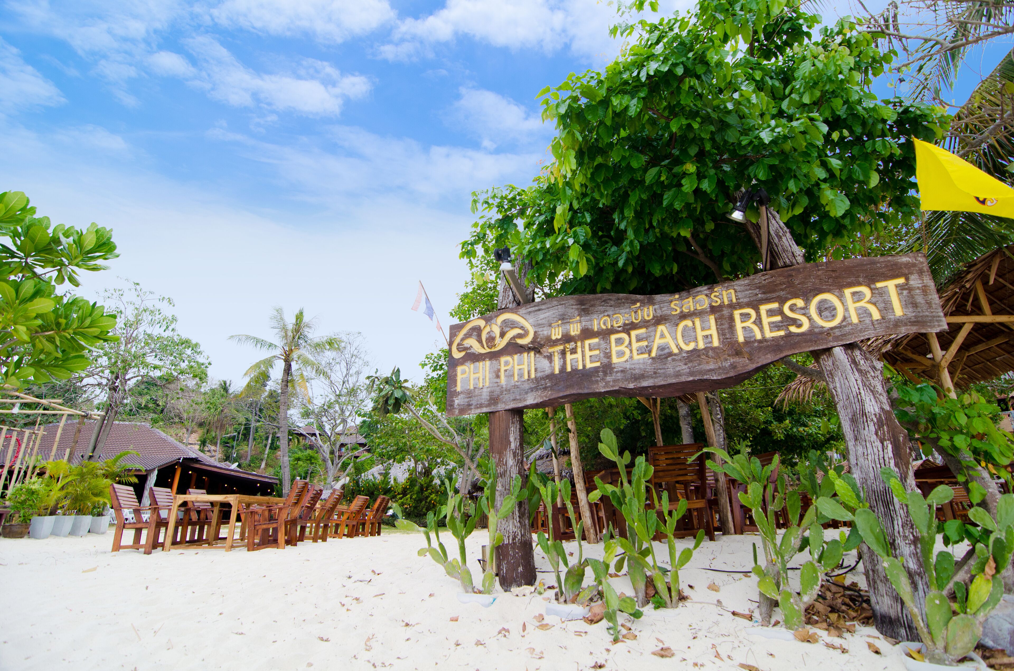 A wooden sign for Phi Phi The Beach Resort with a beach and palm trees in the background.
