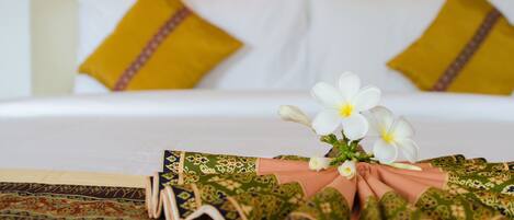Ein ordentlich gemachtes Bett mit weißen und gelben Kissen, einer Blumenkomposition und einer zusammengefalteten Decke mit grün-rosa Muster.