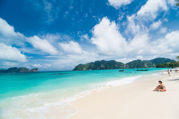 A beach with clear turquoise water, a sandy shore, and a person sitting on the sand.
