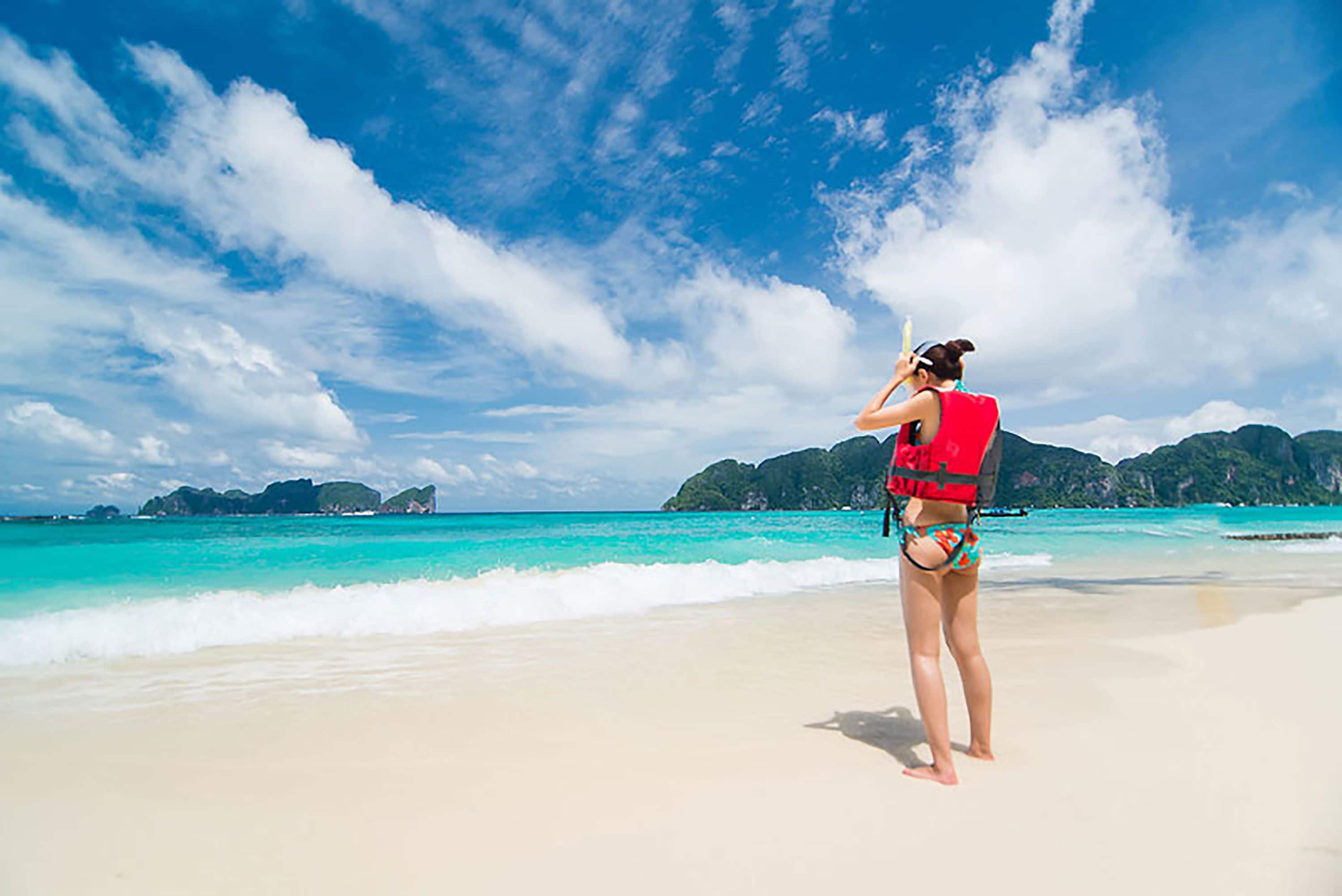 A woman in a red life jacket and bikini on a beach with clear turquoise water and distant green hills.