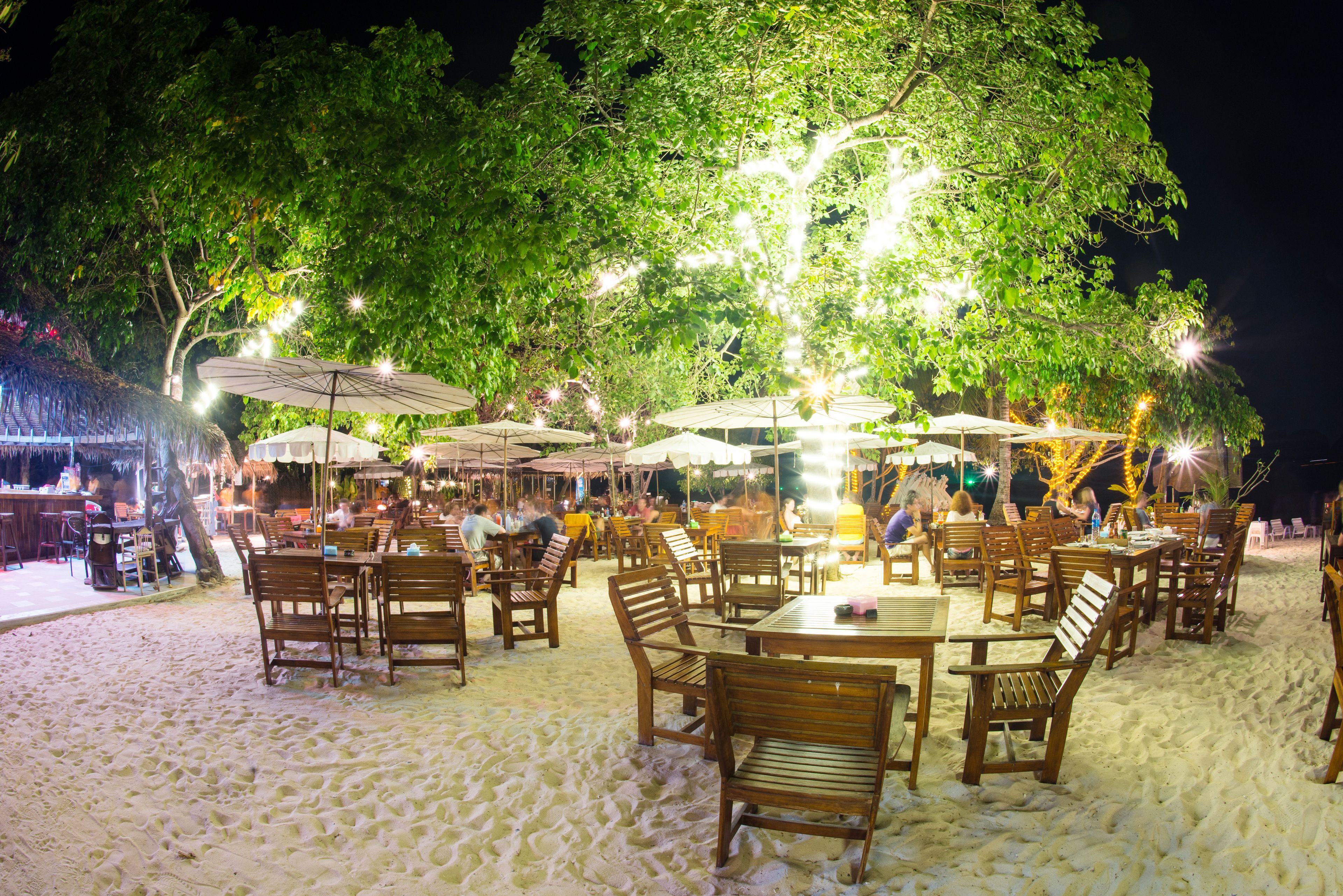 Un espace de restauration en plein air, avec des tables et des chaises, entouré d’arbres et illuminé par des guirlandes lumineuses.
