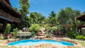 Une piscine agrémentée d’une cascade, entourée d’une végétation luxuriante et d’une structure en bois de style traditionnel.