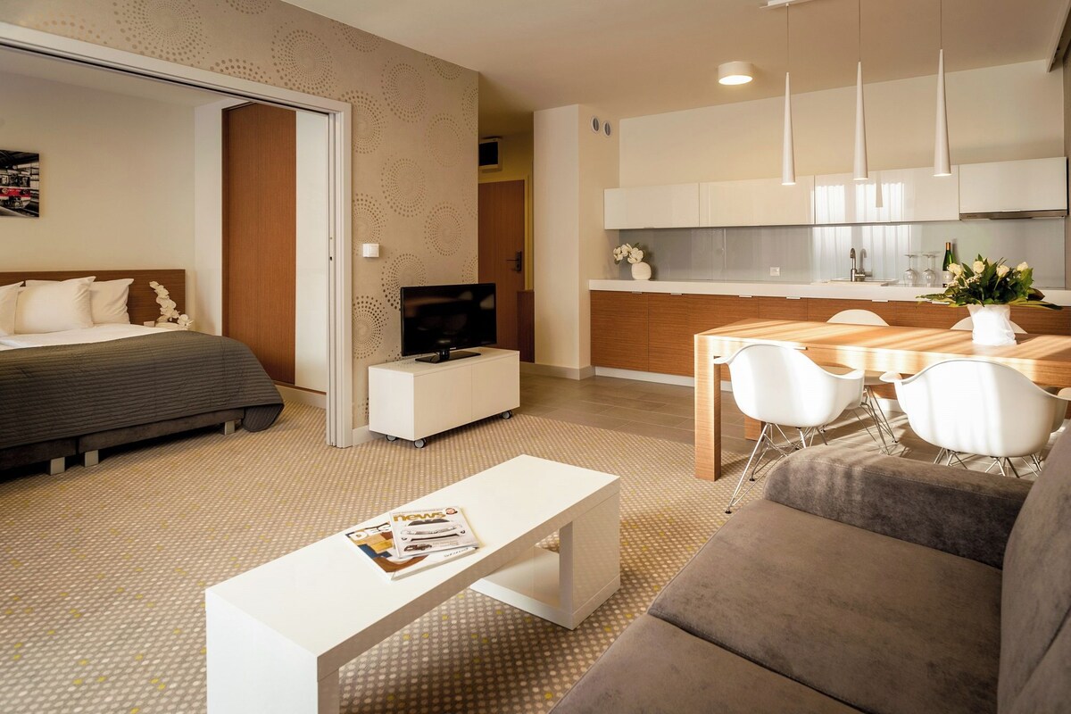 A modern hotel room with a bed, a TV, a dining area with a table and chairs, and a kitchenette.