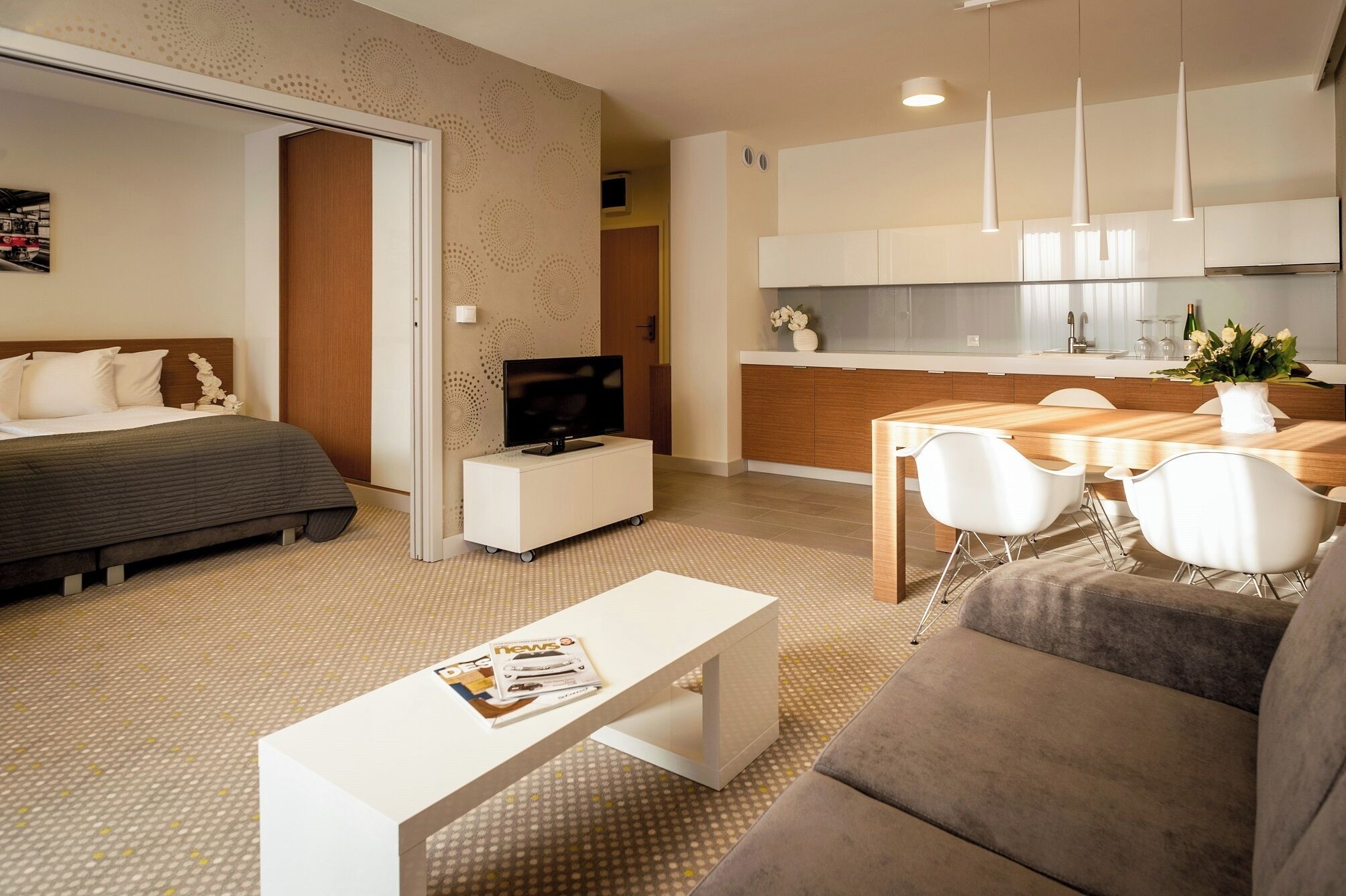 A modern hotel room with a bed, a TV, a dining area with a table and chairs, and a kitchenette.