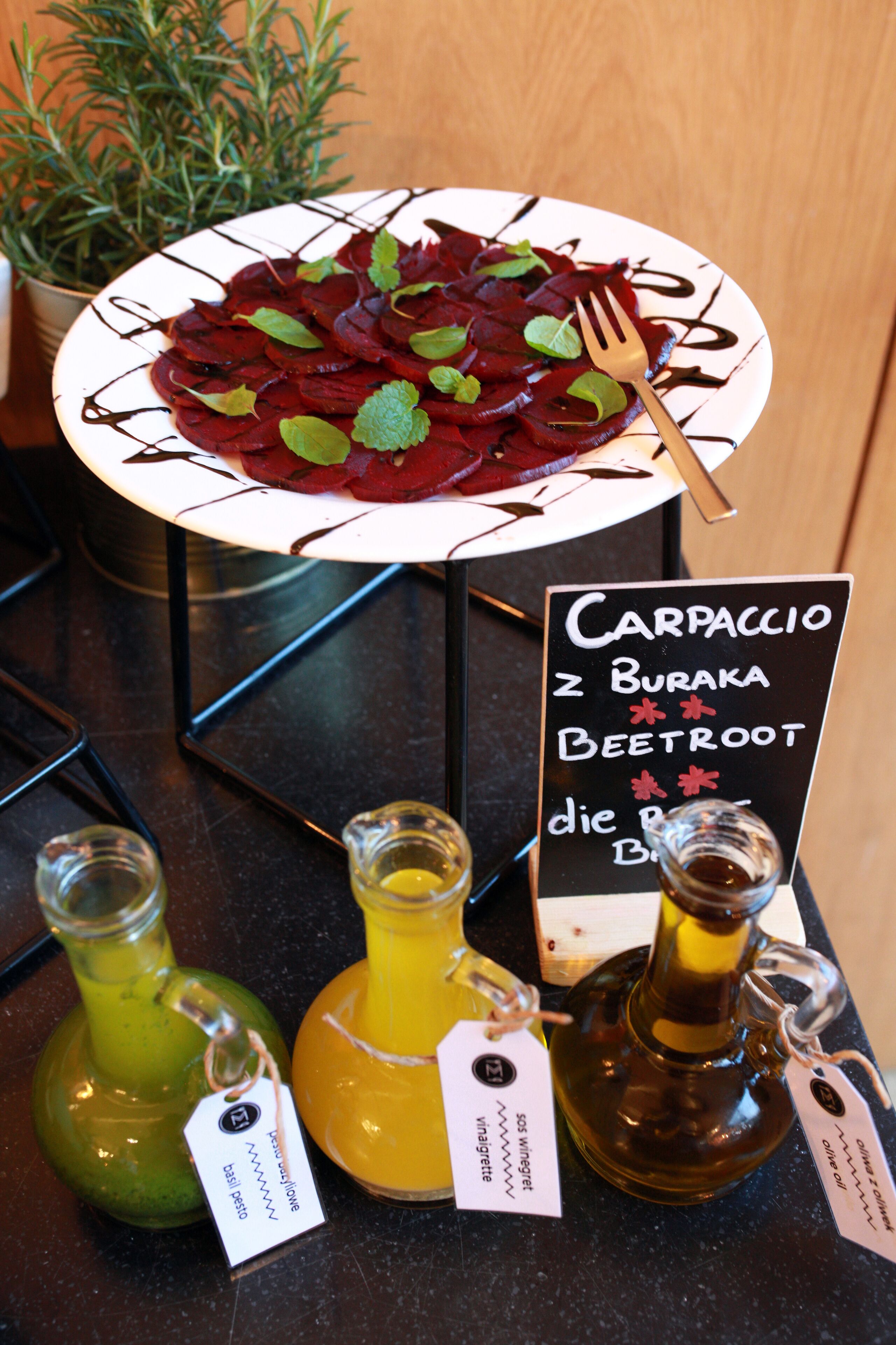 A plate of sliced beetroot salad with a fork, a sign for 'Carpaccio z Buraka Beetroot', and three bottles of olive oil with tags.