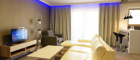 A modern hotel room with a sofa, a TV, a desk, and a dining area.
