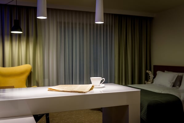 A hotel room with a neatly made bed, a white table with a cup and a newspaper, and a yellow chair.