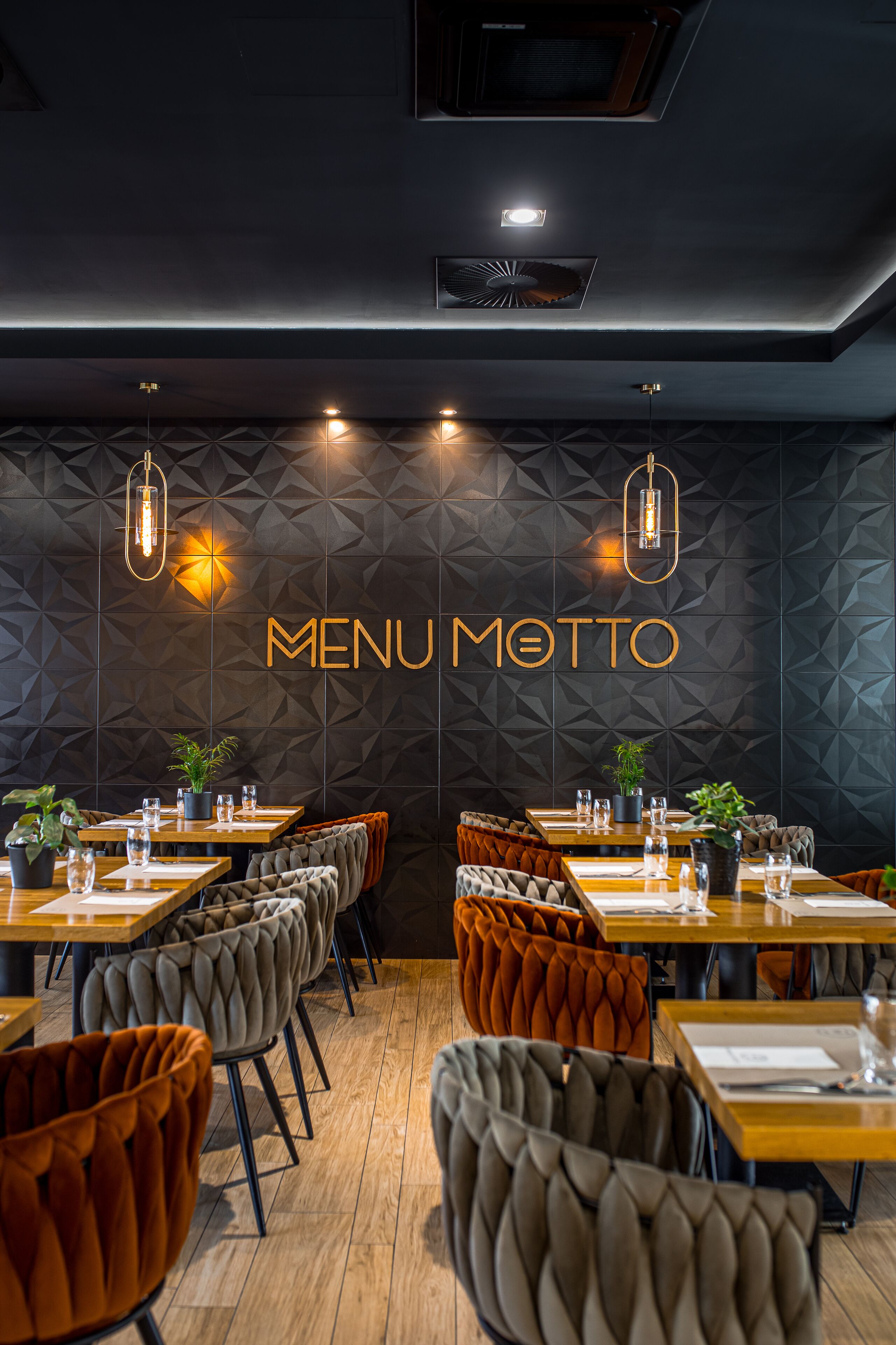 Interior de restaurante con diseño geométrico en la pared, un letrero que dice 'MENU MOTTO', mesas de madera y sillas tapizadas.