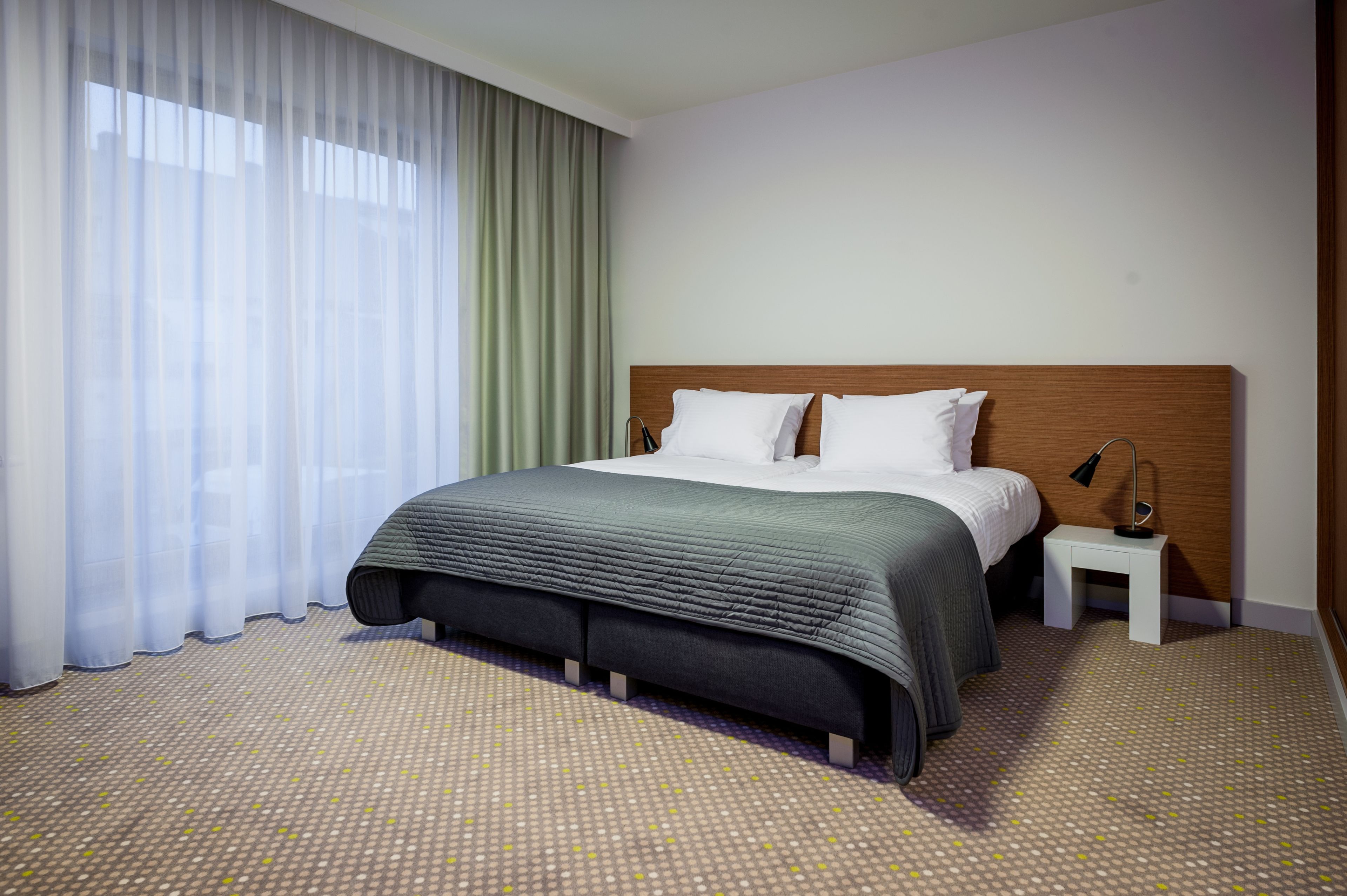 A hotel room with a large bed, a bedside table, a lamp, and sheer curtains.