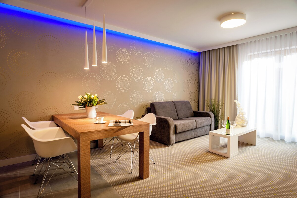 A modern living room with a wooden dining table, white chairs, a grey sofa, and a small white side table with a bottle and glass.