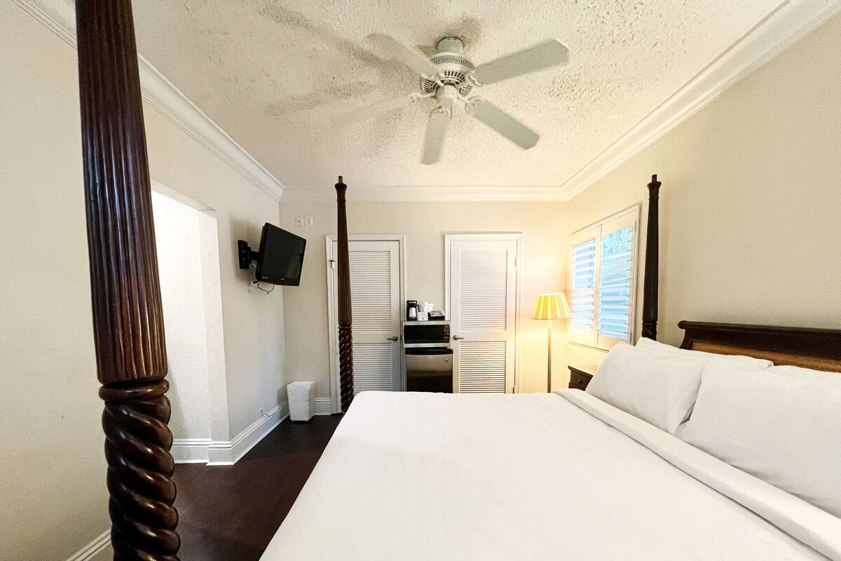 A hotel room with a large bed, a ceiling fan, a television mounted on the wall, a desk with a lamp, and a window with blinds.