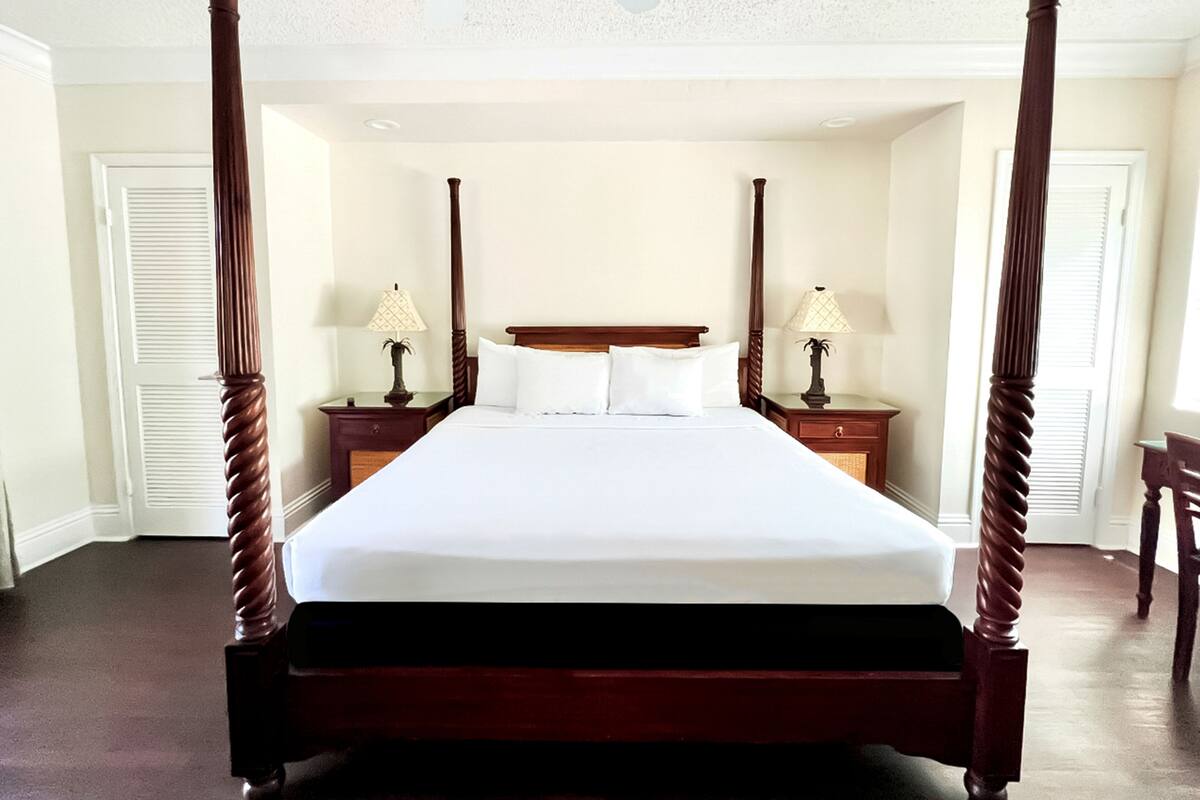A four-poster bed with white linens, two bedside tables with lamps, and a ceiling fan.