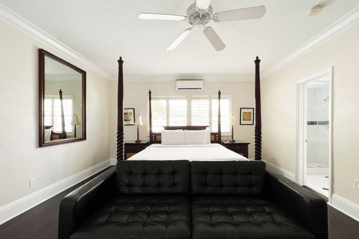 A four-poster bed with a ceiling fan, a large mirror, and a dark upholstered sofa in a hotel room.