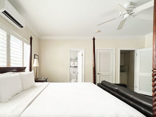 Habitación de hotel con una cama grande, ventilador de techo, ventana con persianas y puerta que da a un baño.
