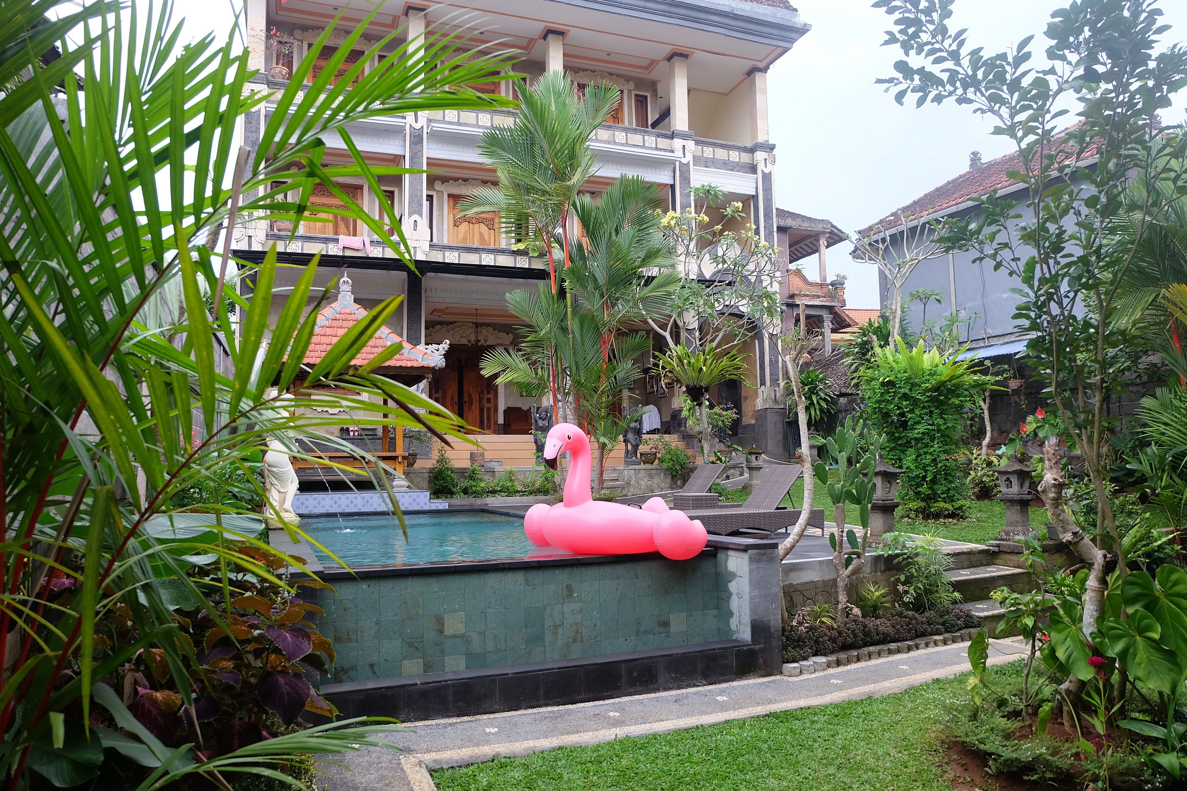A pool with a pink flamingo float, surrounded by lush greenery and a multi-story building with balconies.