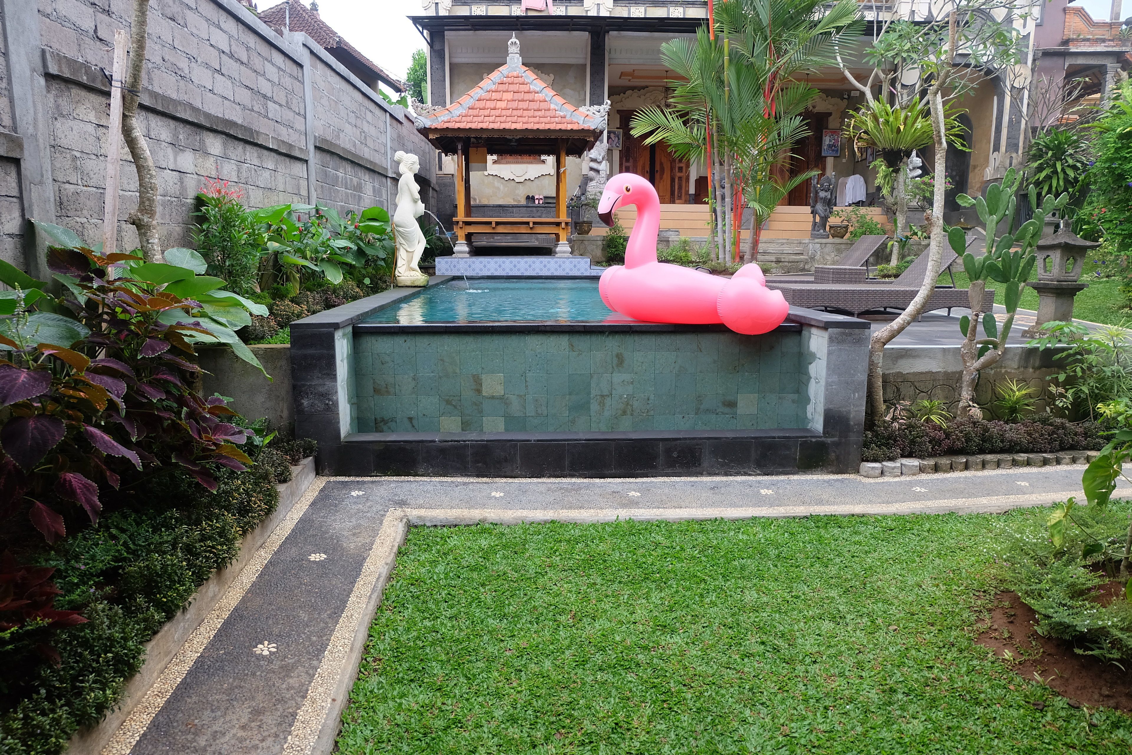 En swimmingpool med en lyserød flamingo-svømmebøje, omgivet af grønt og en traditionel sommerpavillon.