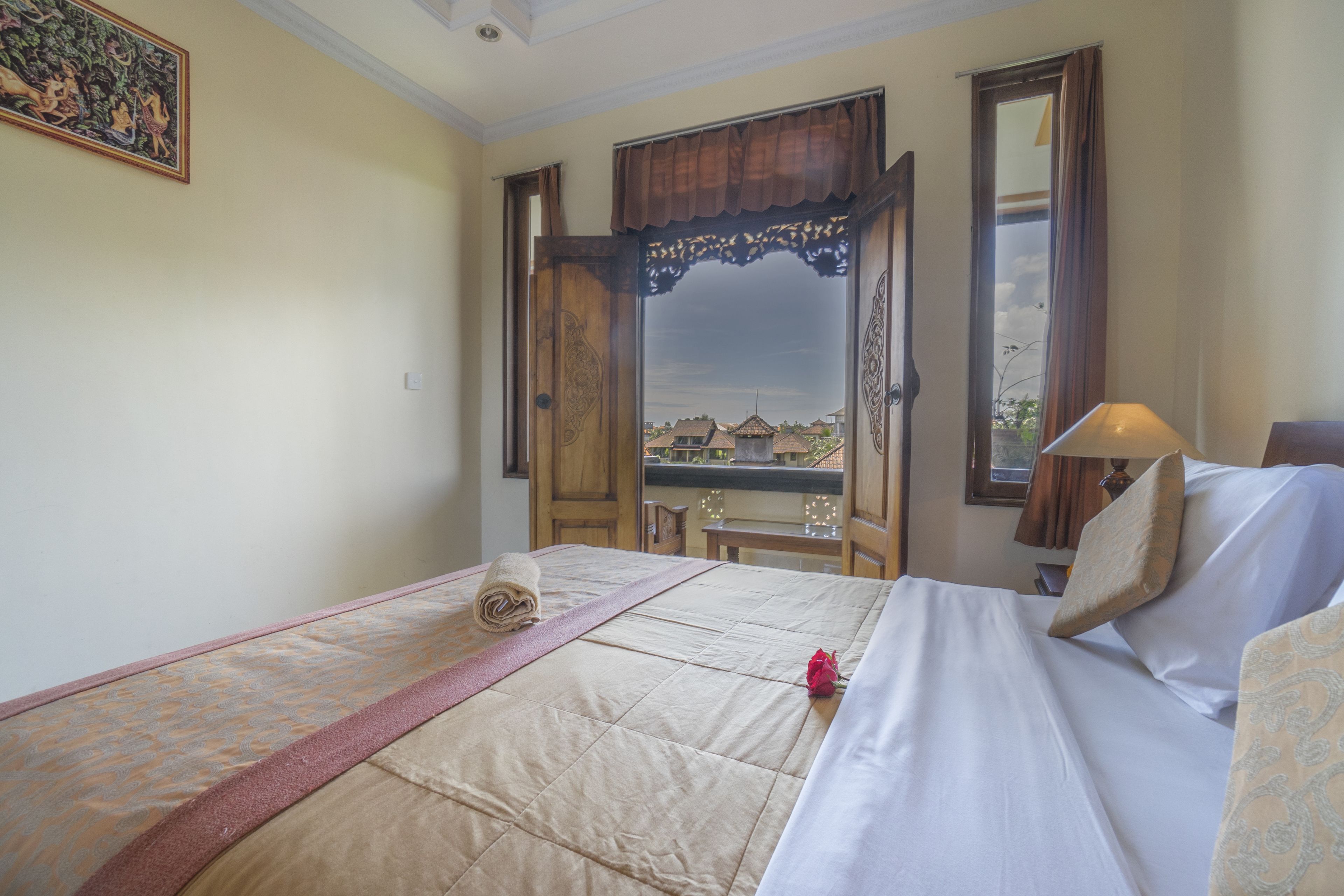A hotel room with a bed, a window with a view of buildings, a wooden door, and a painting on the wall.