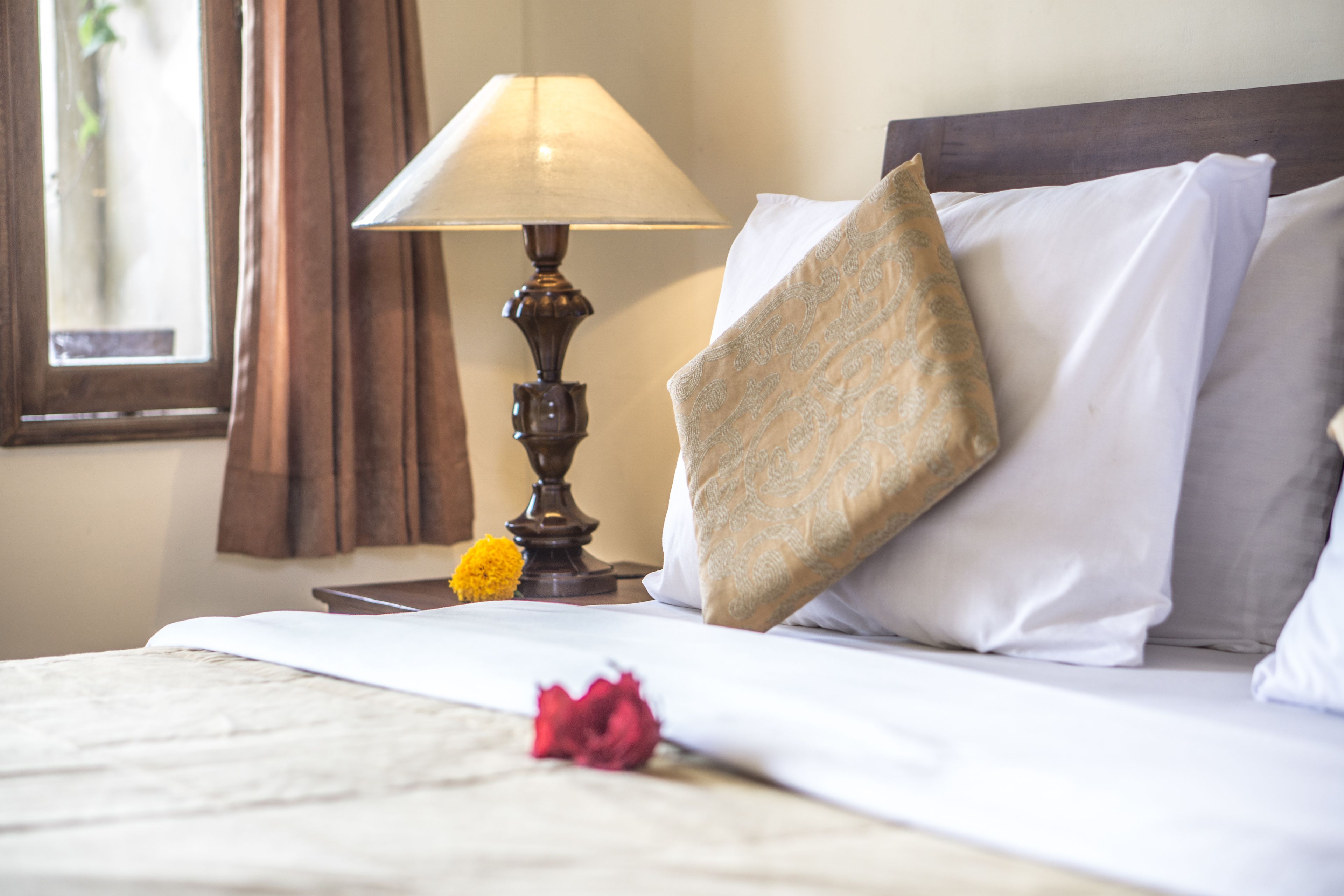 Un lit bien fait, avec du linge de lit blanc, un coussin décoratif, une lampe de chevet et une fleur posée sur le lit.