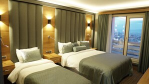 Habitación de hotel con dos camas, un ventanal grande con vistas a la ciudad y un cabecero de madera con iluminación integrada.