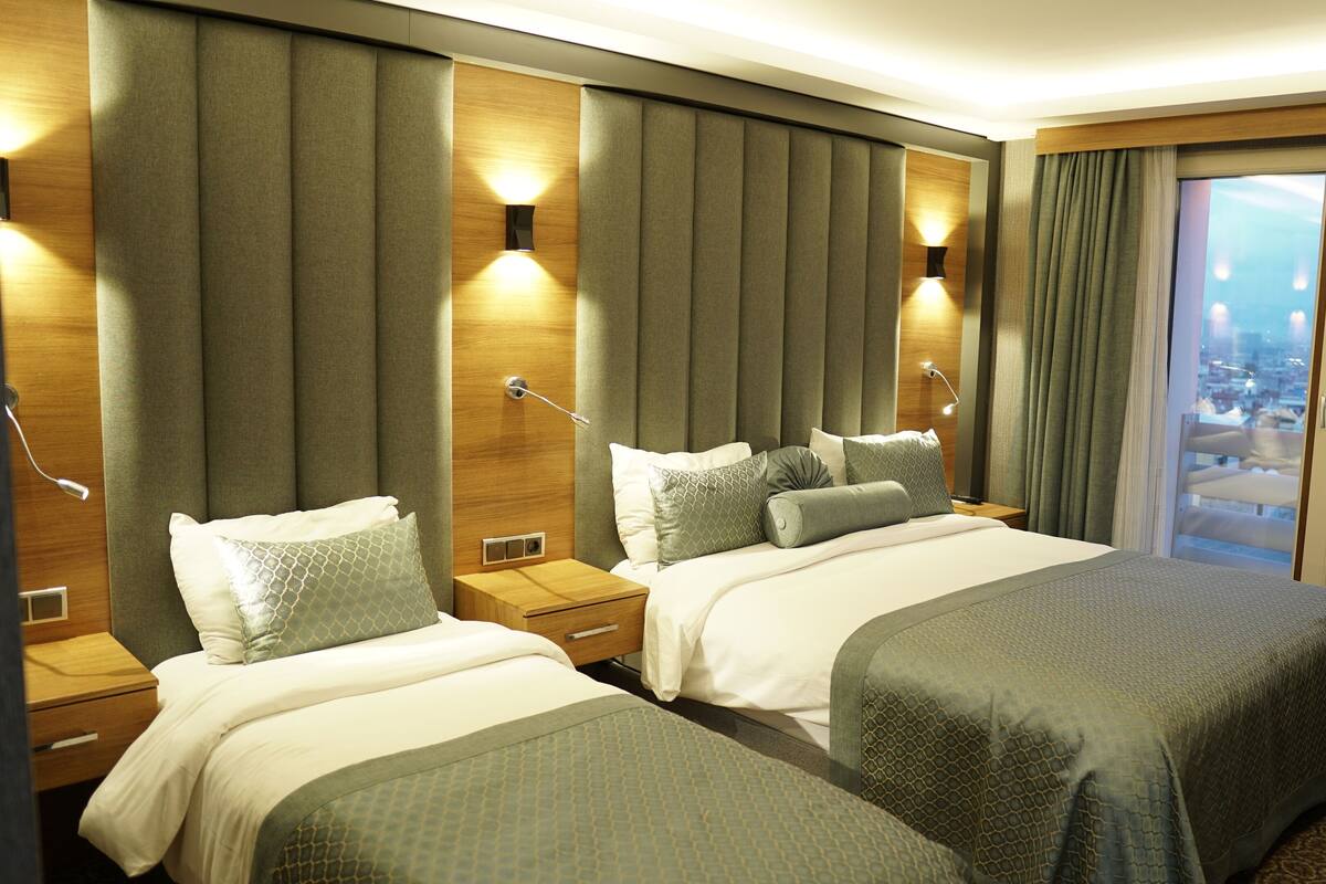 A hotel room with two beds, a large window with a city view, and a wooden headboard with built-in lighting.
